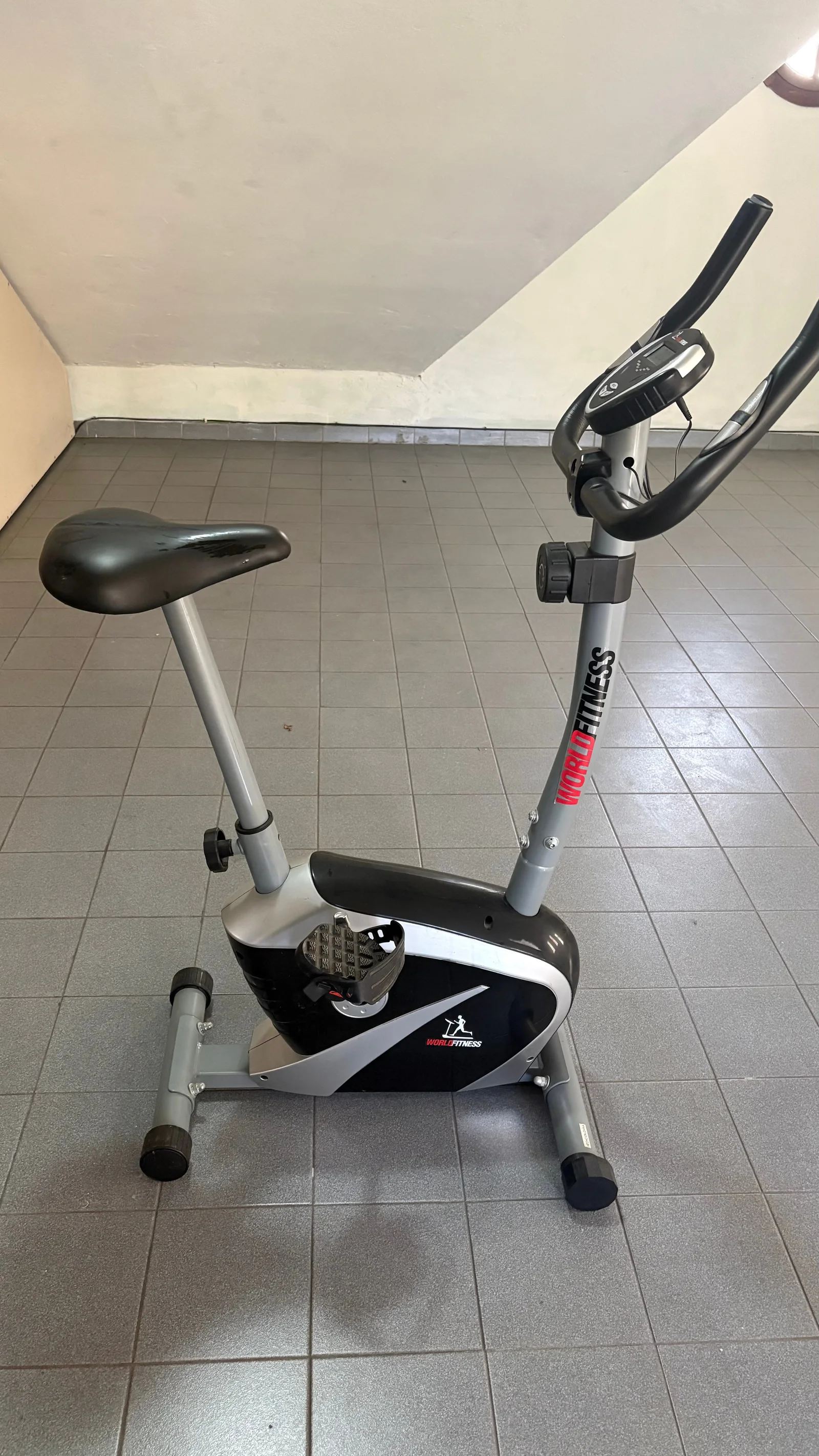 Bicicleta fija World Fitness gris y negra vista de costado, con asiento, pedales y consola