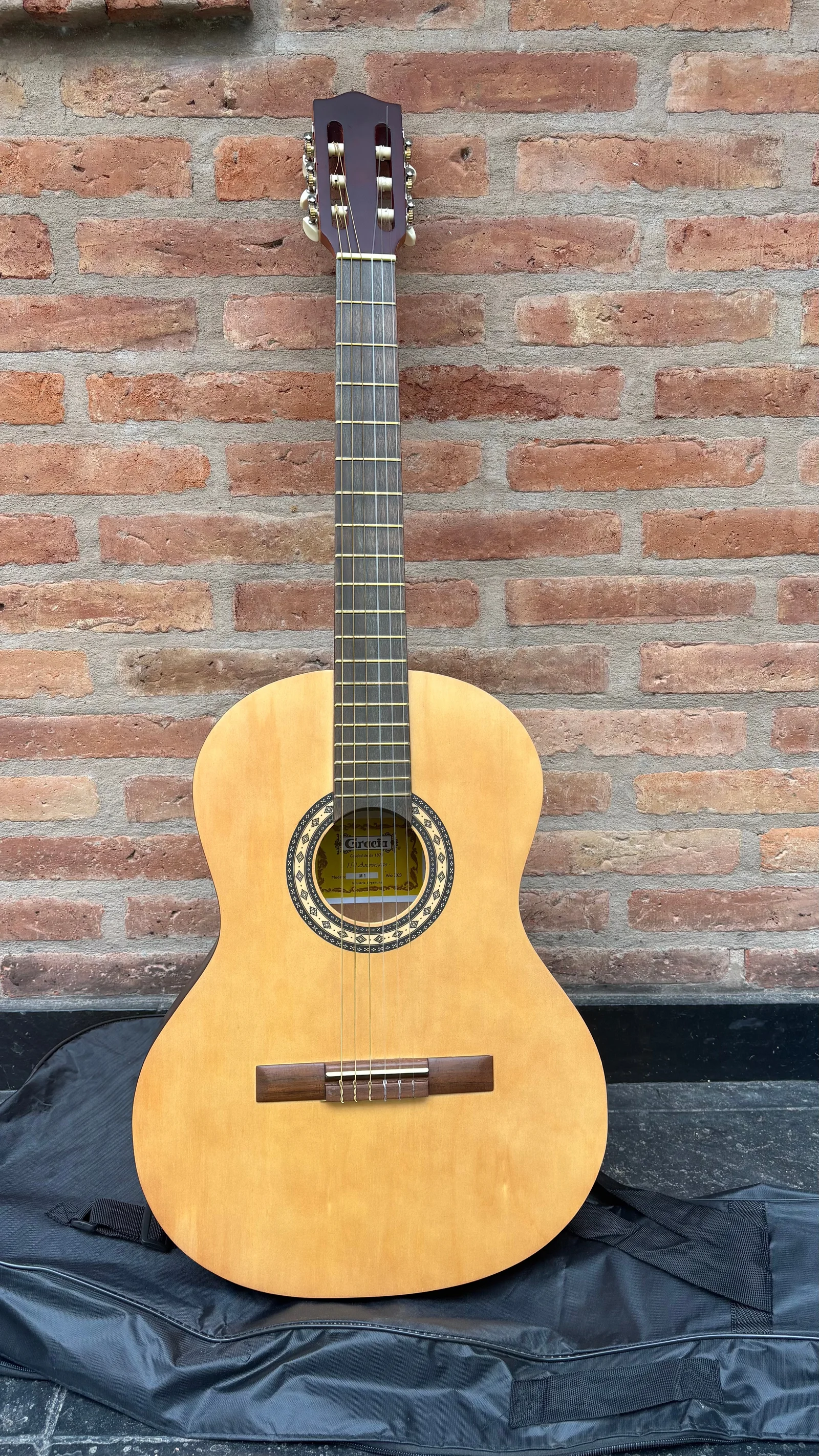 Guitarra criolla Gracia vista de frente, apoyada sobre su funda contra una pared de ladrillos